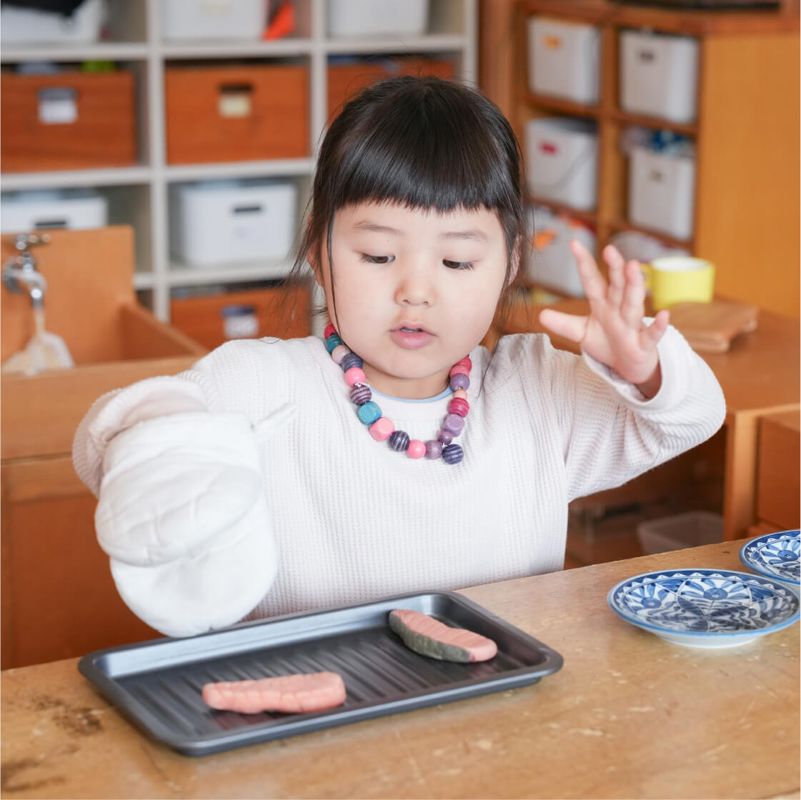 白いミトンをはめた手で、鉄板に載せた粘土のようなものを焼く真似をしておままごとを楽しむ女の子。