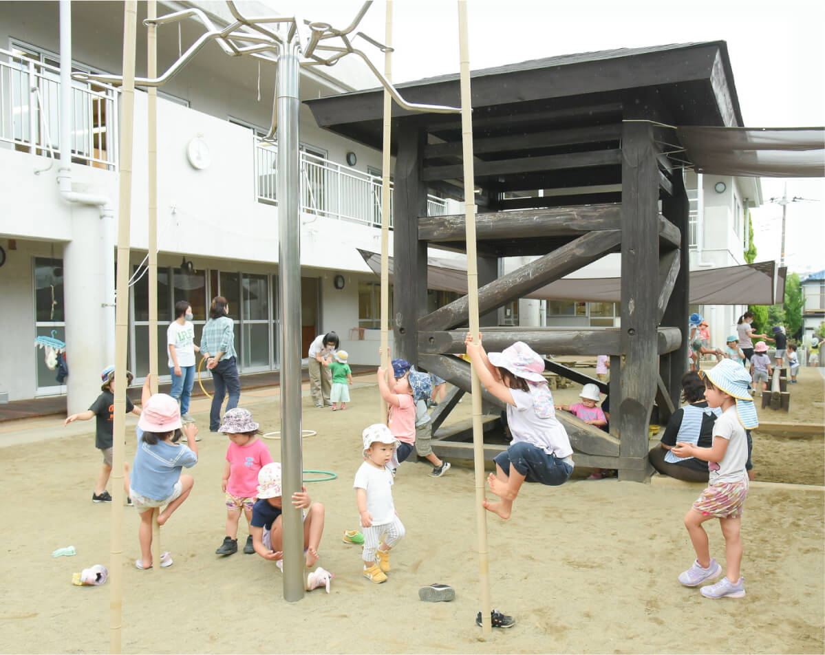 園庭の砂場にある木製のやぐらや金属製の登り棒などの遊具で、元気に遊ぶたくさんの子どもたち。