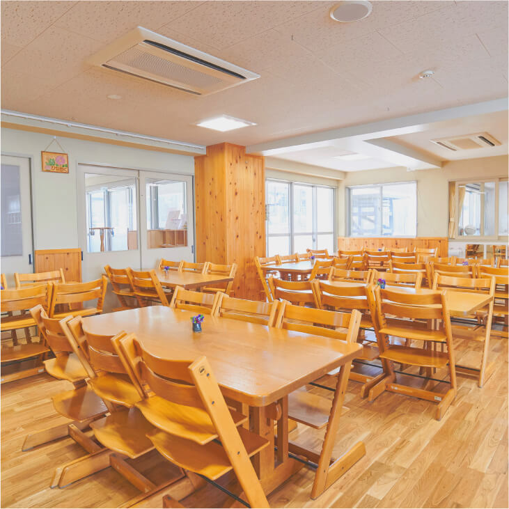 食事や作業に使われる、木製のテーブルと子ども用の椅子が整然と並べられた明るい保育室。