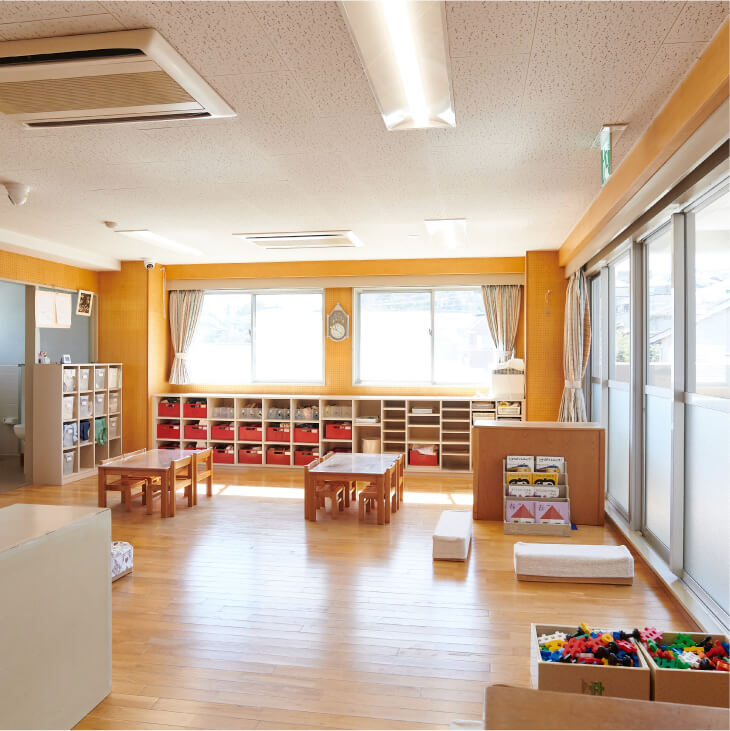 壁に沿ってたくさんのおもちゃや道具が整理された木製の棚が並ぶ保育室。中央には小さなテーブルと椅子がある。
