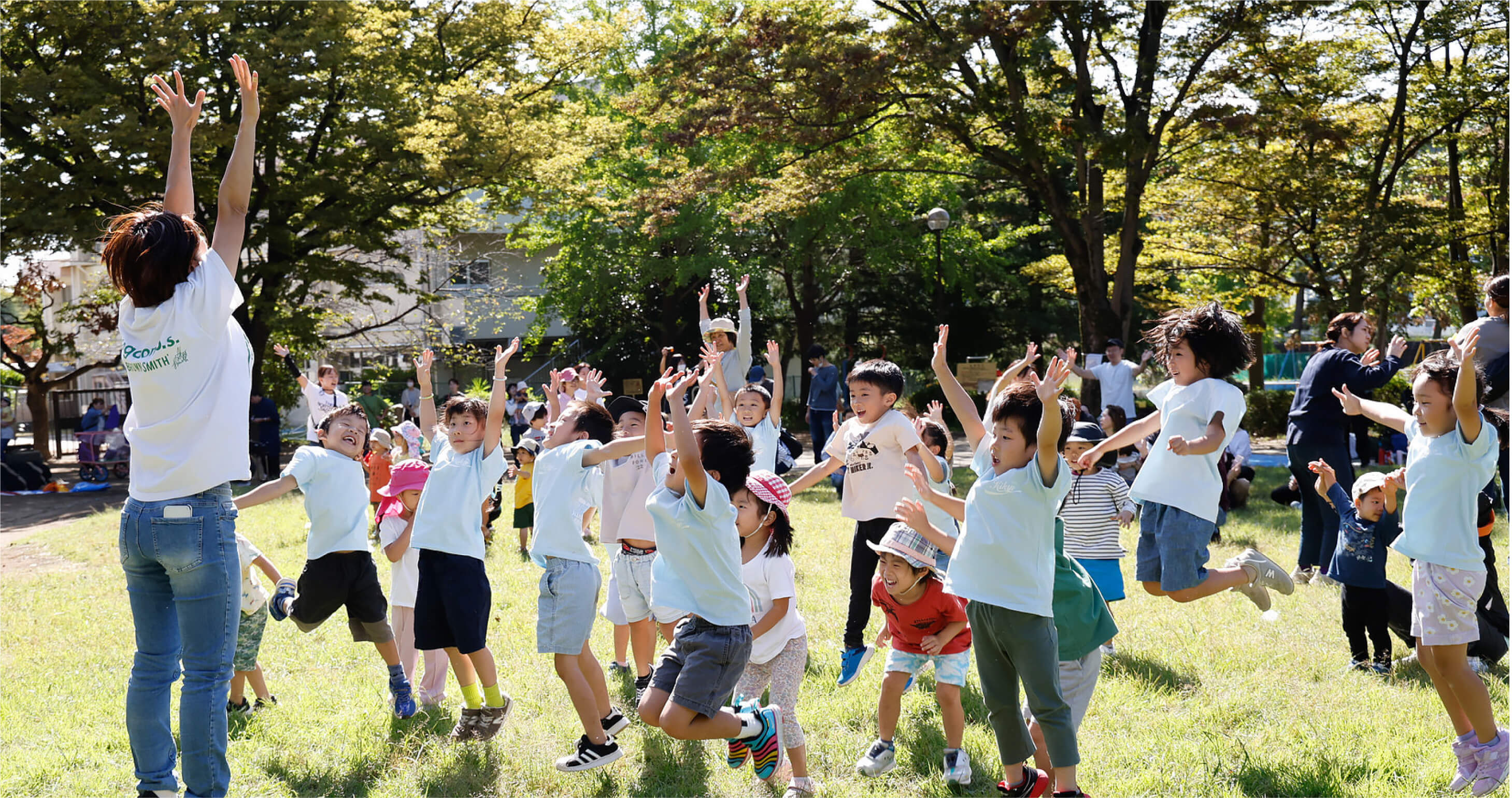 緑豊かな芝生の公園で、職員も子どもたちも一緒になって両手を高く挙げ、元気いっぱいにジャンプする様子