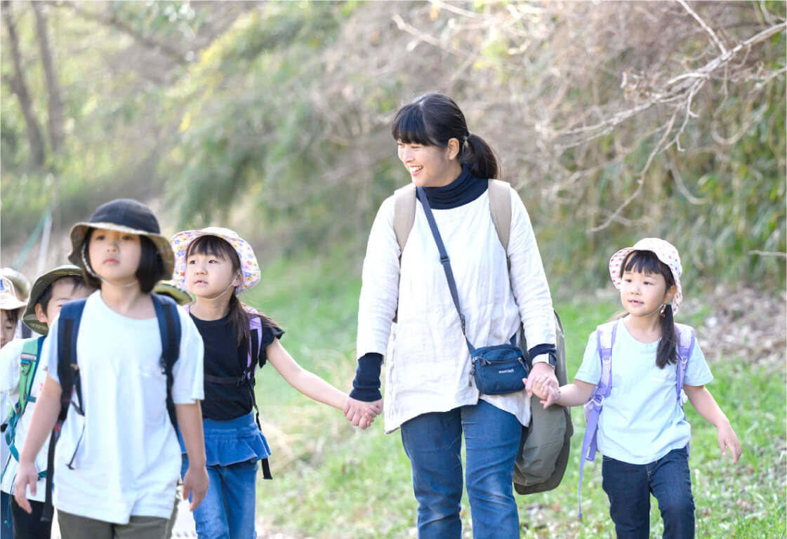 子どもたちと手をつないで自然豊かな道を歩く保育士