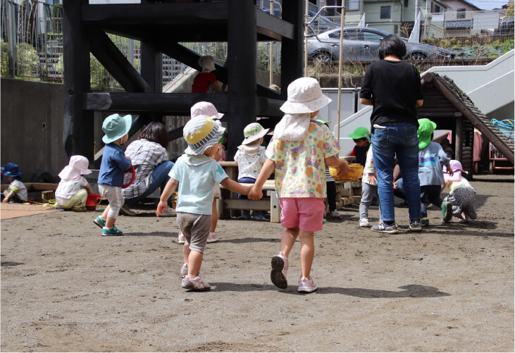 園庭で仲良く手を繋いで歩く、年齢の異なる子どもたちの後ろ姿