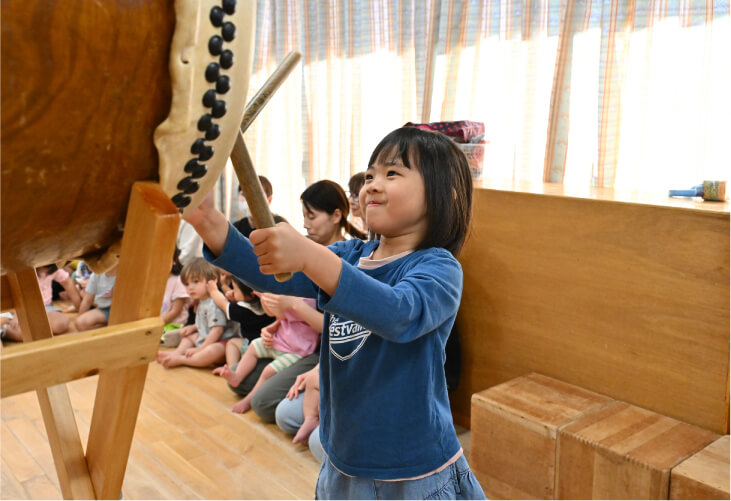 お友達が見守る中、室内で大きな和太鼓をバチで力強く叩く女の子