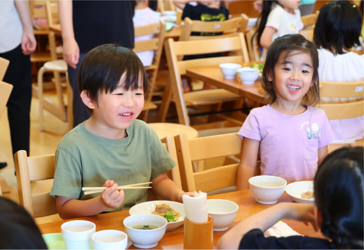明るいランチルームの木製テーブルで、お友達と楽しそうに給食を食べる子どもたち