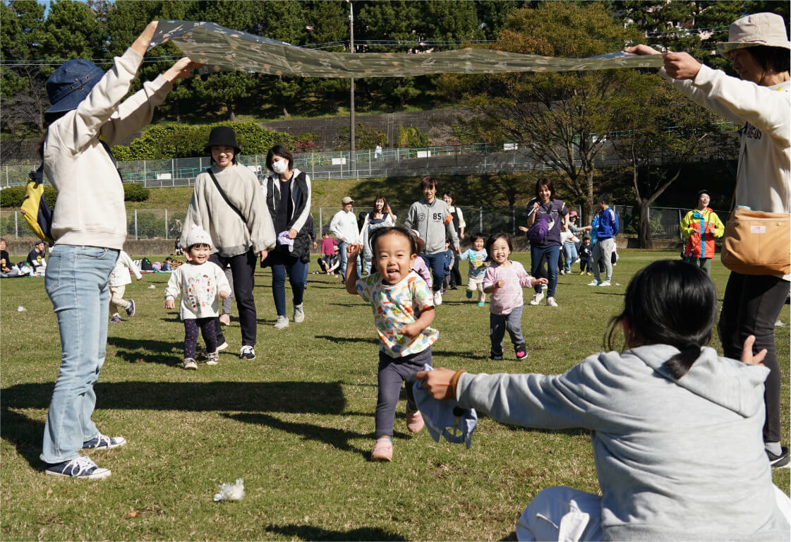 緑豊かな広い公園で、先生と一緒に両手を挙げて楽しそうに遊ぶ子どもたち