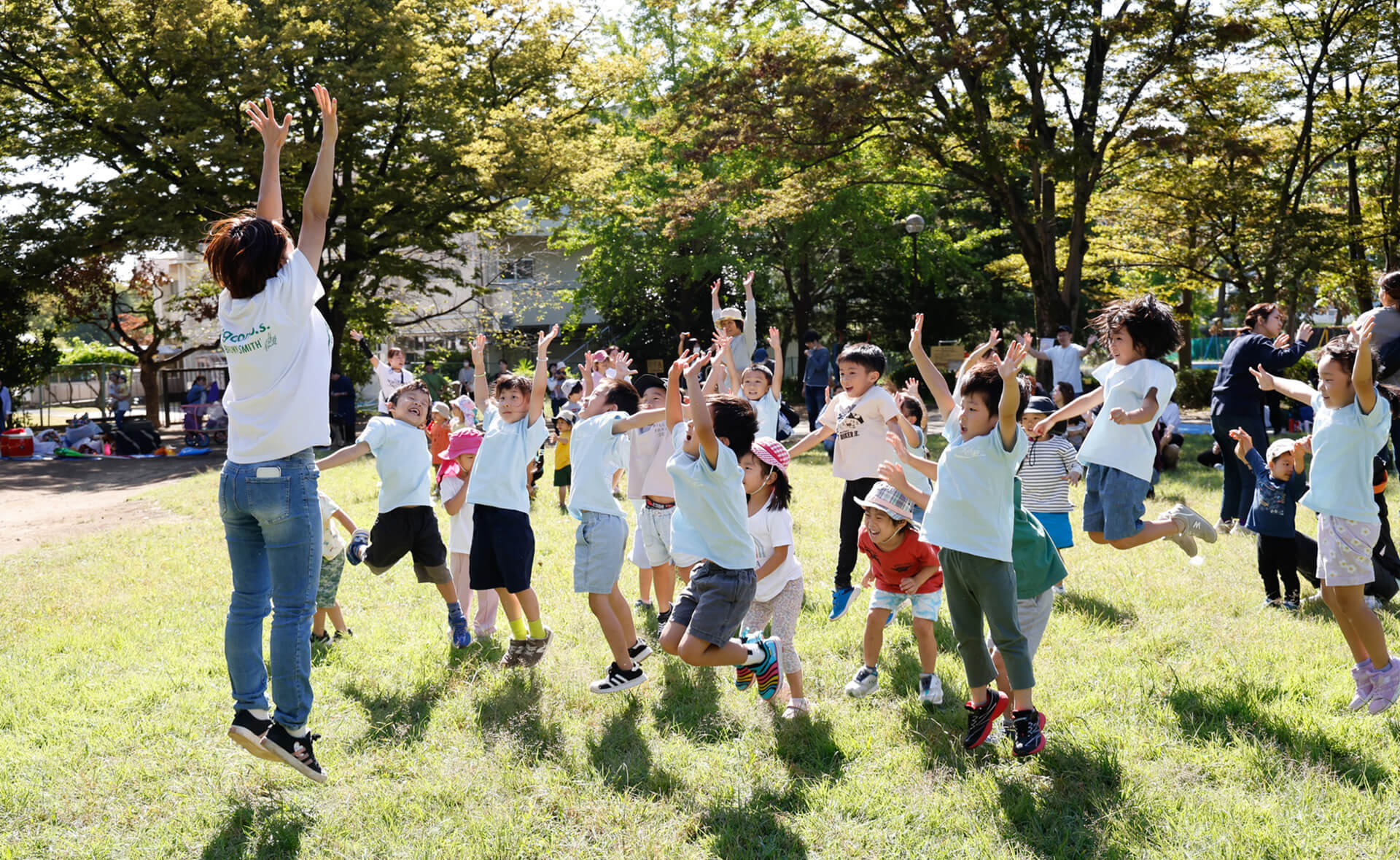 緑豊かな芝生の公園で、職員も子どもたちも一緒になって両手を高く挙げ、元気いっぱいにジャンプする様子