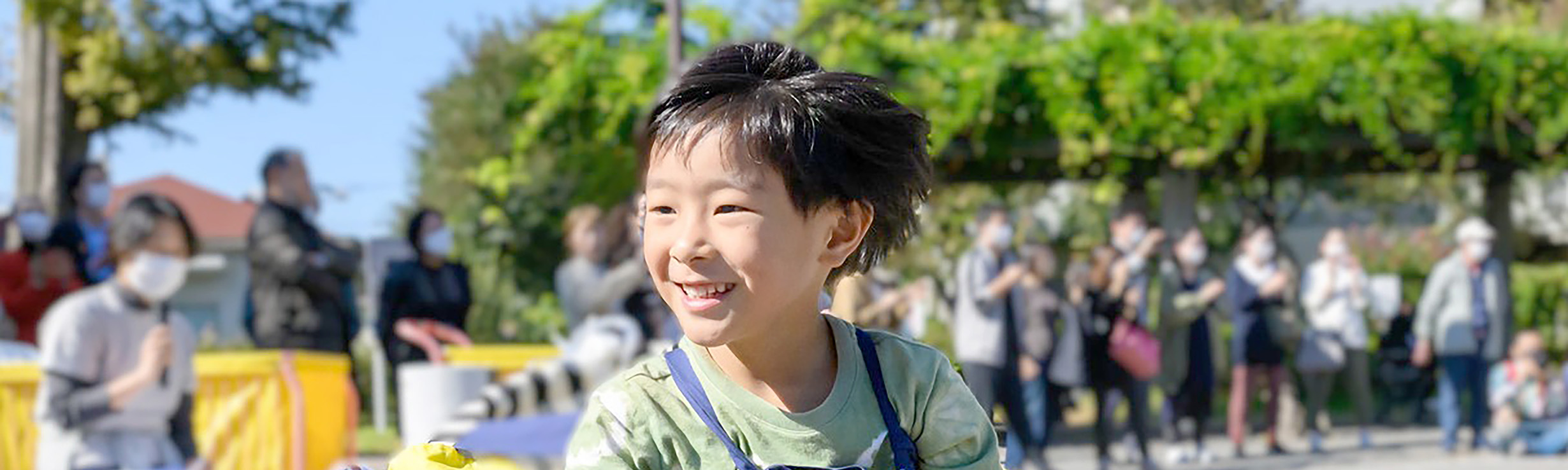 園庭で緑色のシャツを着て馬のような飾りを身につけ、笑顔で元気に走る男の子。背景には見学する保護者たちの姿がある。