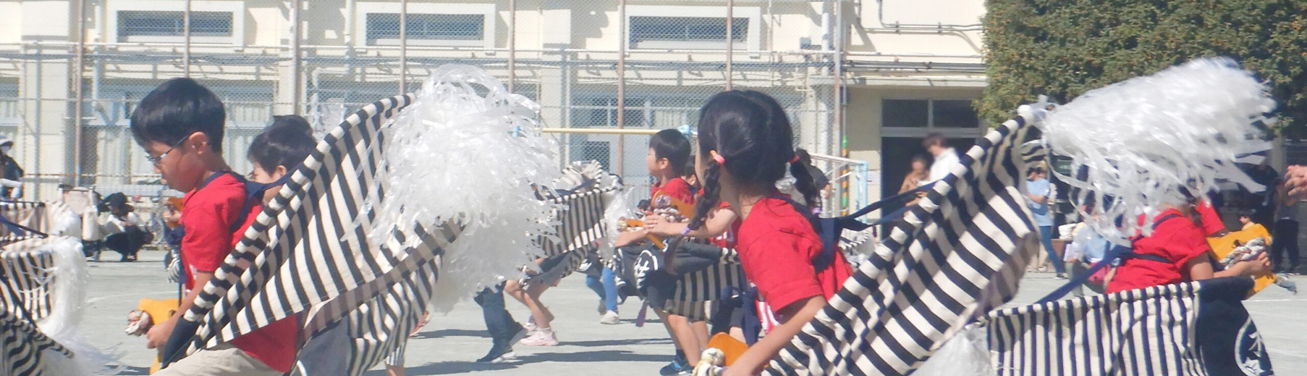 赤いTシャツを着て白黒の縞模様の馬の飾りを身につけ、園庭で元気に踊る子どもたち。