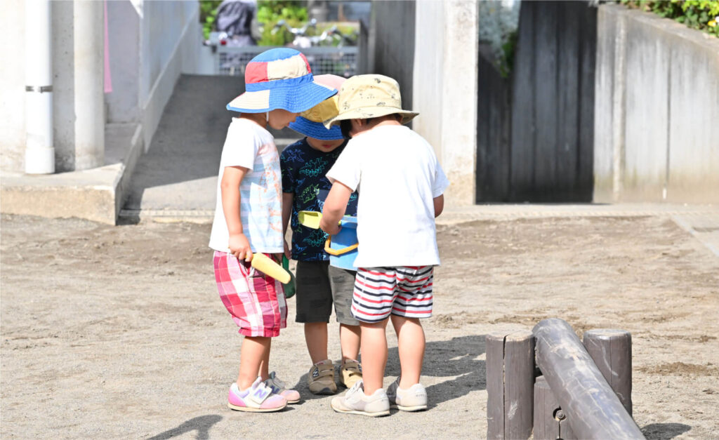 砂場で、真ん中の男の子を囲んで話し合うか、何かを見つめる、帽子を被った3人の男の子。手前には丸太の遊具がある。
