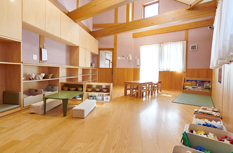 木材をふんだんに使った明るく広々とした保育室。おもちゃが整理された棚、緑色のローテーブルとクッション、奥には小さな机と椅子が配置されている。