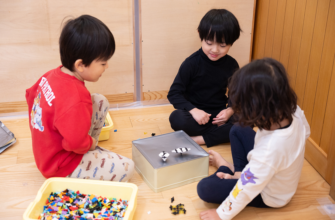 木の床に座ってブロックのおもちゃで遊ぶ3人の子どもたち。裏返した四角い缶の上で、ブロックで作ったコマを回して楽しんでいる様子。