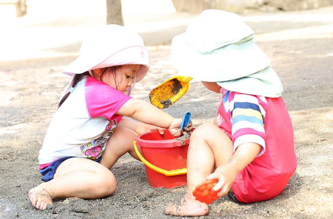 日よけ帽子と水着を身につけ、はだしで泥んこ遊びをする2人の幼児。黄色いスコップを使って赤いバケツに湿った砂を入れている。