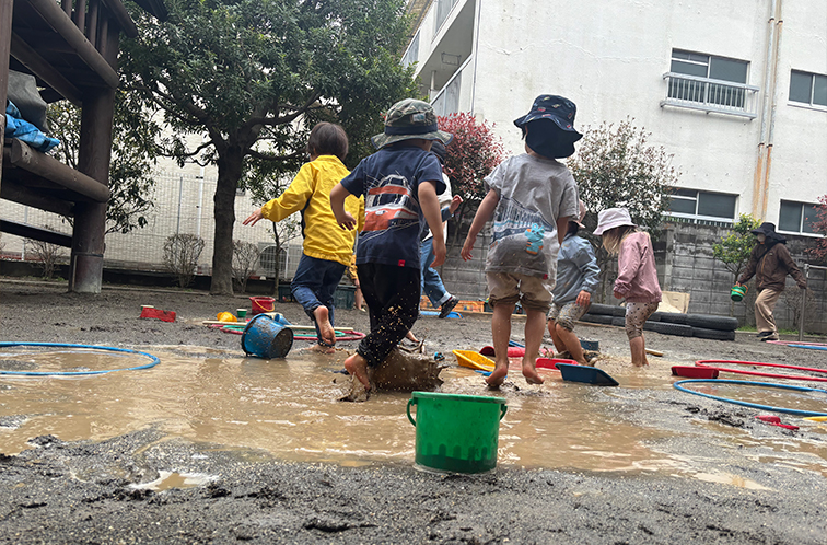 園庭の大きな水たまりで泥んこ遊びをする子どもたち。はだしで泥水に飛び込み、バケツやフラフープなどの遊具とともに全身で遊んでいる様子。