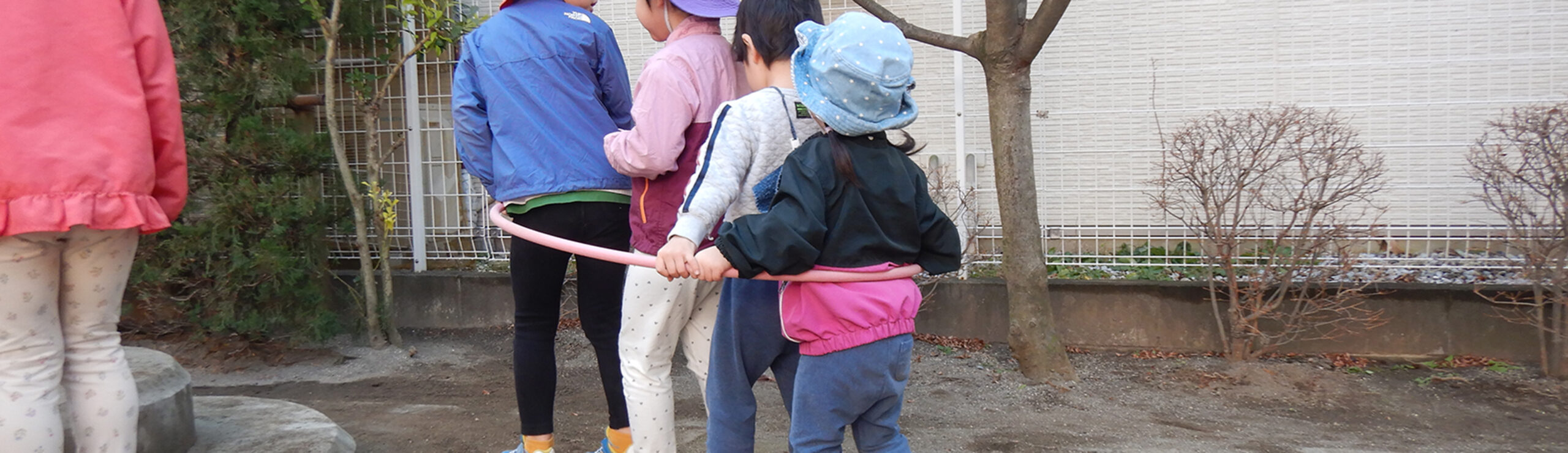 園庭で一つのピンク色のフラフープの中に複数人で入り、電車ごっこのように連なって遊ぶ子どもたちの後ろ姿。