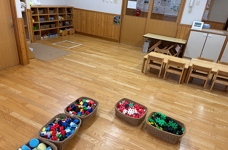 木の床が広がる広々とした保育室。手前には色鮮やかなブロックのおもちゃが入ったカゴが並び、奥には子ども用の小さな木製テーブルと椅子、壁際に収納棚が配置されている。